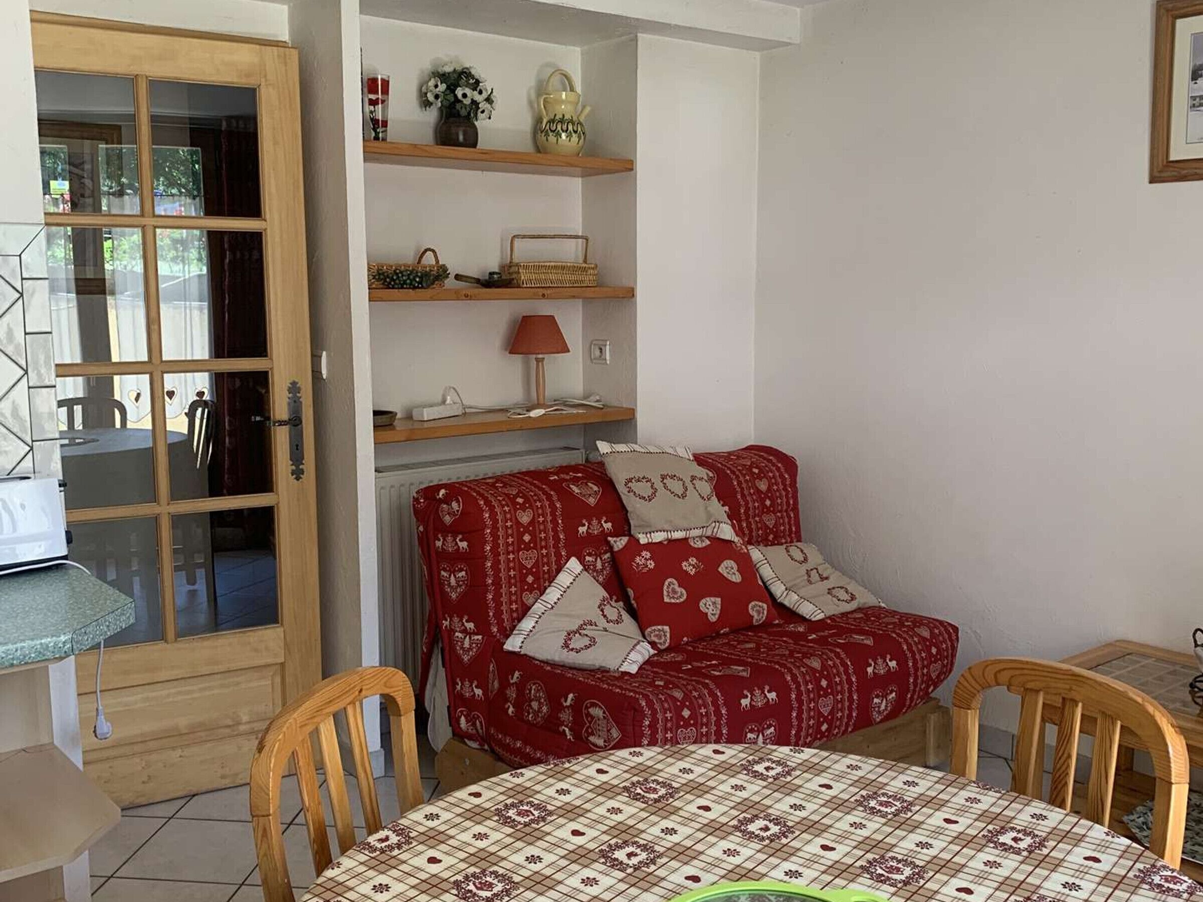 Et hyggeligt rum med en trædør, en rød sofa med hjertepude, et lille bord med dug og en reol med pynteting.