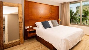 Una camera da letto con una testiera in legno, un grande specchio, un letto con biancheria da letto bianca e vista su palme attraverso una finestra.