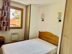 A bedroom with a wooden bed, a window with curtains, a wall-mounted heater, and a view of a wooden building outside. - Cozy apartment - 4 guests (Valloire)
