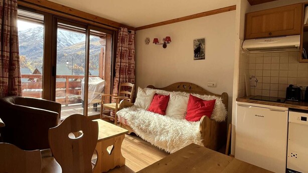 Una stanza accogliente con un tavolo da pranzo in legno, un divano con cuscini rossi e bianchi, un angolo cottura con un frigorifero bianco e vista sulle montagne attraverso ampie finestre.