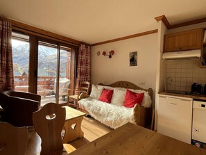 A cozy room with a wooden dining table, a sofa with red and white cushions, a kitchen area with a white refrigerator, and a view of mountains through large windows. - Apartment with balcony - 4 guests (Valloire)