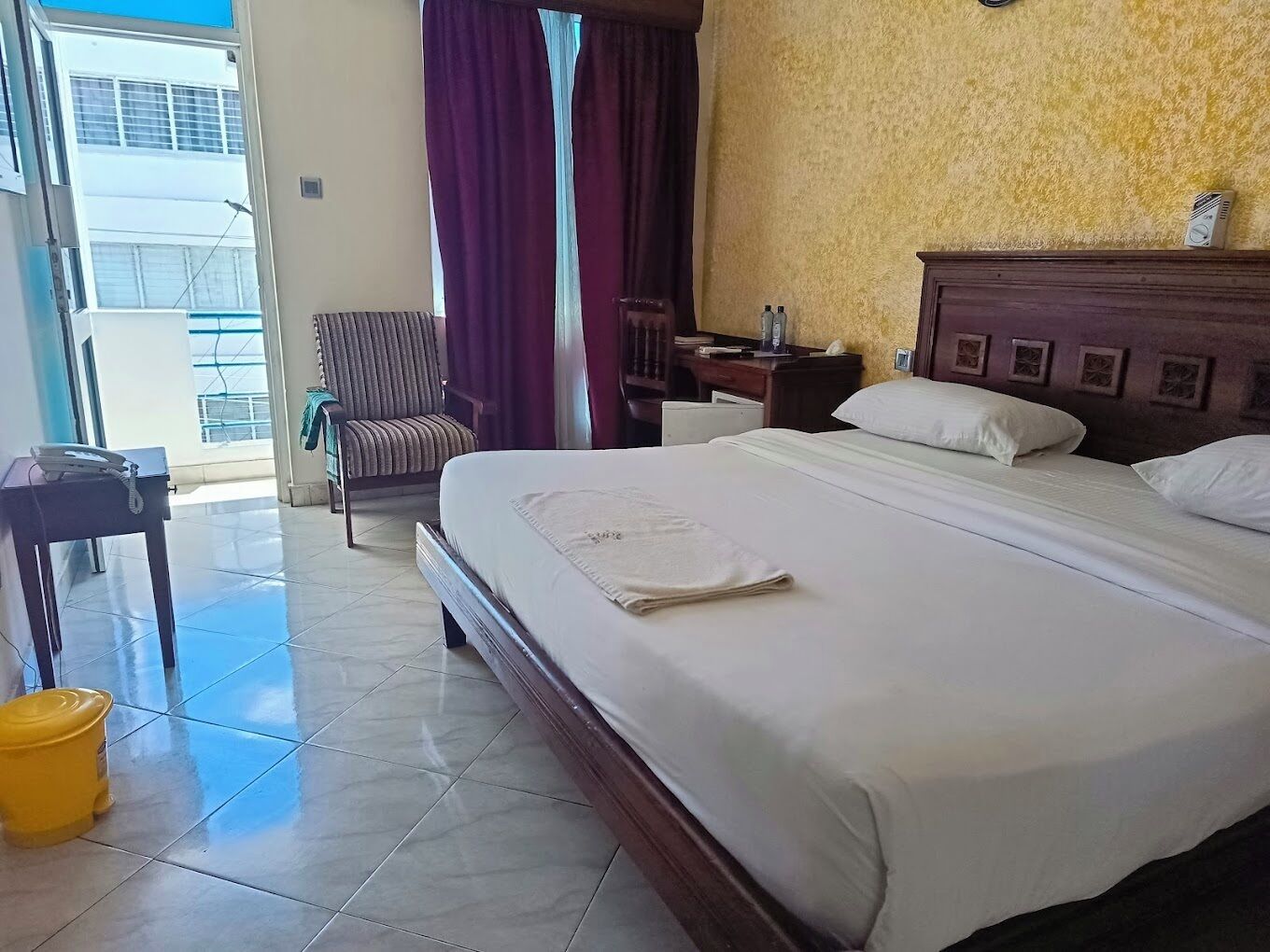 A hotel room with a large bed, a chair, a desk with a telephone, and a view of the outside.