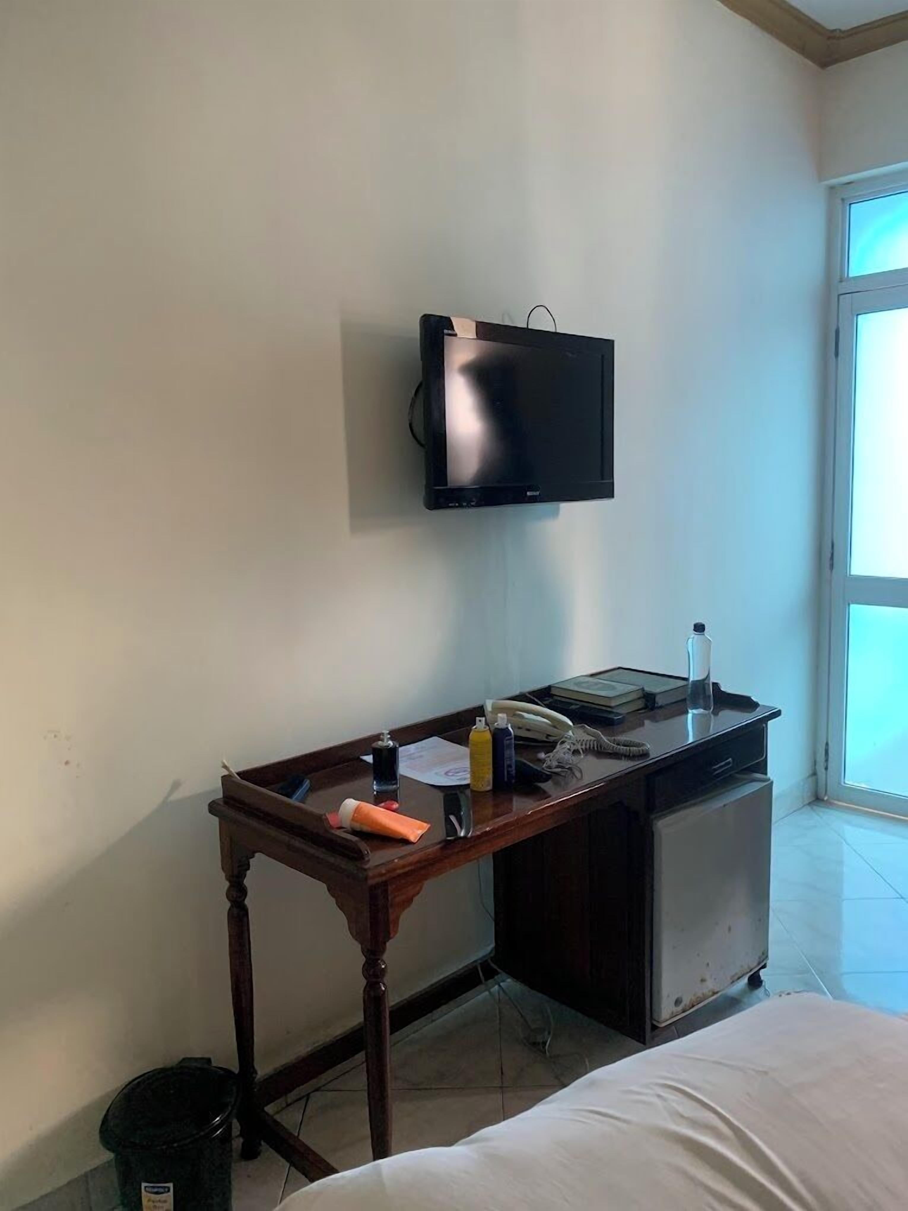 A hotel room with a desk, a television mounted on the wall, a bed, and a window.