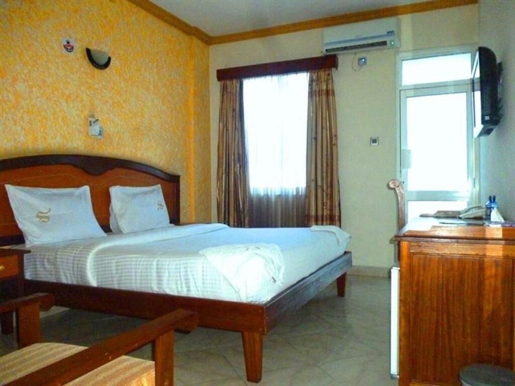 A hotel room with a wooden bed, a desk, a chair, a window with curtains, and an air conditioning unit.