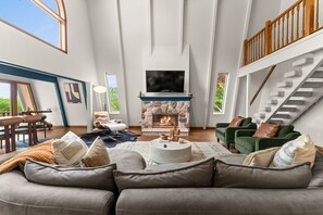 A spacious living room with a stone fireplace, a large TV mounted above it, and a staircase with wooden railings.