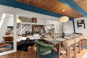 A spacious loft with a high ceiling, a dining area with a large table and chairs, a staircase, and a cozy seating area with sofas and a coffee table.