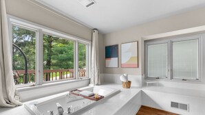 Un baño moderno con una bañera grande, una ventana con vista a árboles y una terraza de madera afuera.