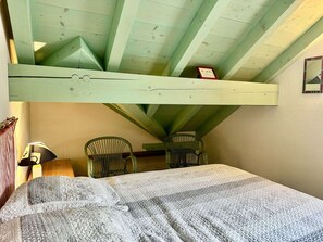 Een kamer met een schuine plafond, twee groene stoelen, een bed met een grijze deken en een klein lijstwerk met een afbeelding aan de muur.