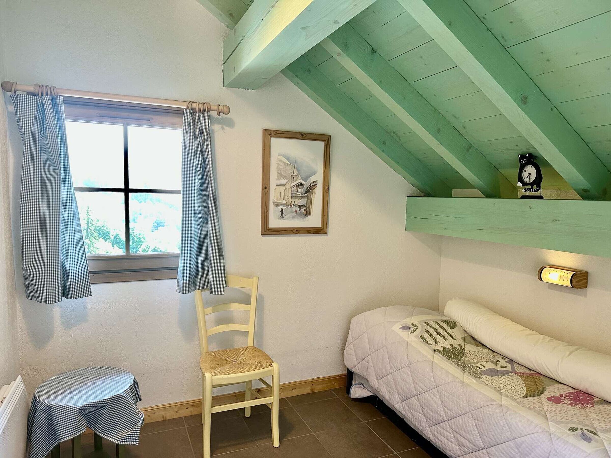 Un dormitorio con cama, una silla, una mesita, una ventana con cortinas, un cuadro enmarcado y un reloj en la pared.