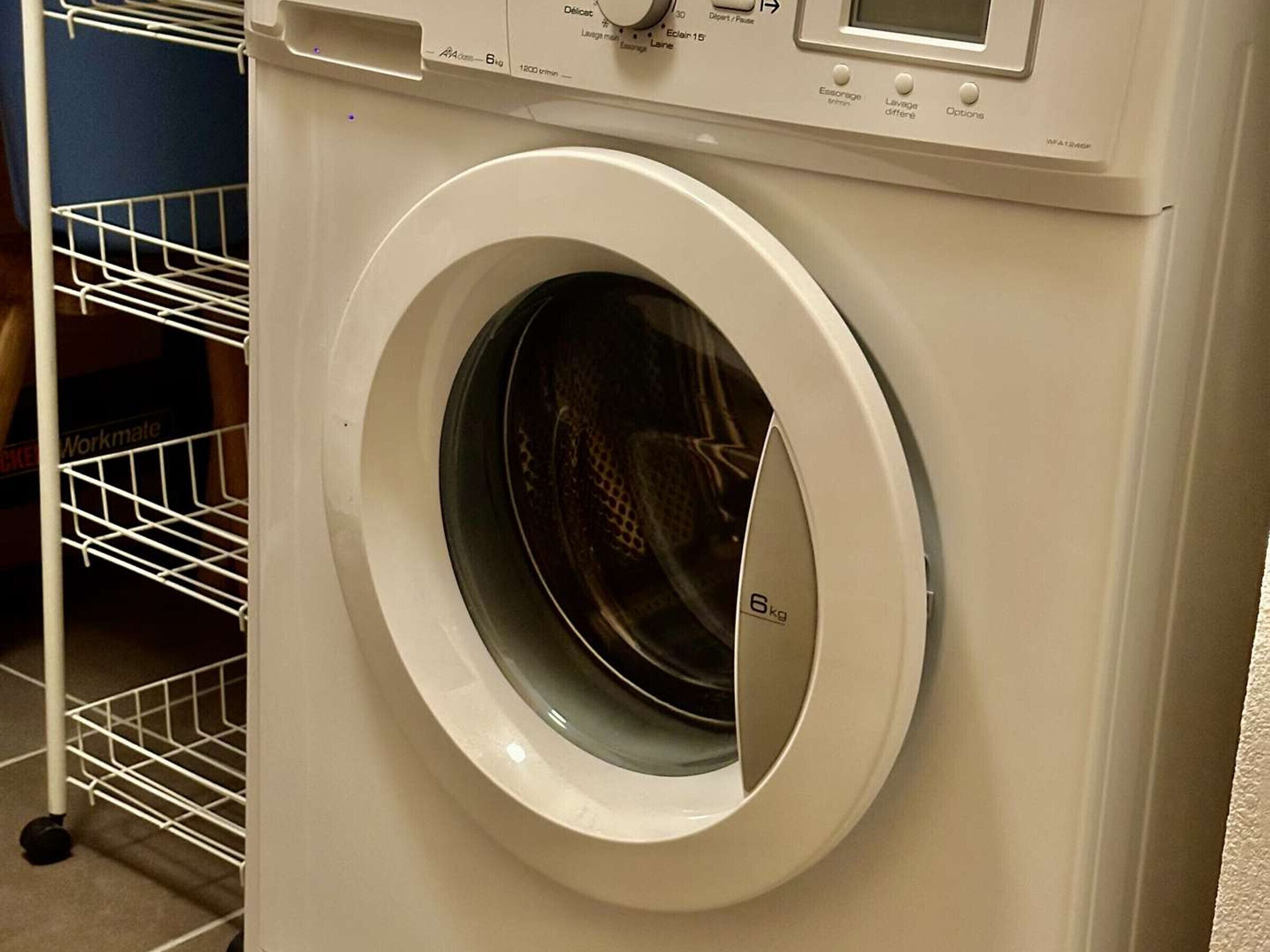 A front view of a front-loading washing machine with a control panel and a metal shelf to its left.