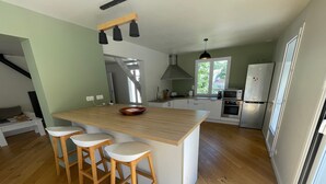 A modern kitchen with a central island, wooden stools, stainless steel appliances, and a large window providing natural light. - Villa in the pines near the beach, parking and pets allowed (La Tranche-sur-Mer)