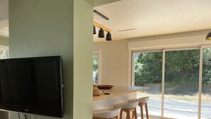 A modern kitchen with a large TV, wooden stools, and a sliding glass door leading to a garden. - Villa in the pines near the beach, parking and pets allowed (La Tranche-sur-Mer)