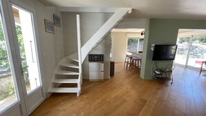 A spacious room with a wooden floor, a white staircase, a kitchen area with a bar, and a television mounted on the wall. - Villa in the pines near the beach, parking and pets allowed (La Tranche-sur-Mer)