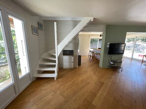 A spacious room with a wooden floor, a white staircase, a kitchen area with a bar, and a television mounted on the wall. - Villa in the pines near the beach, parking and pets allowed (La Tranche-sur-Mer)