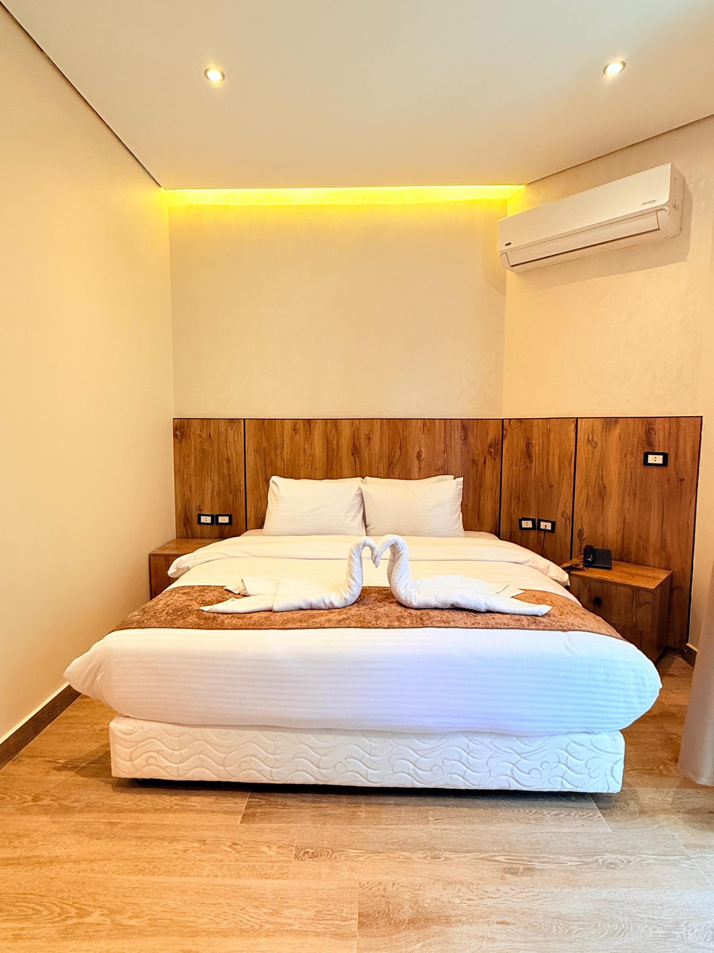 A modern hotel room with a wooden headboard, a neatly made bed, a bedside table, and an air conditioning unit.