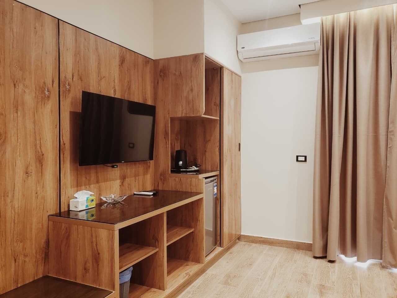 A compact room with a wooden wardrobe, a flat-screen TV, a small kitchenette, and a sliding door.