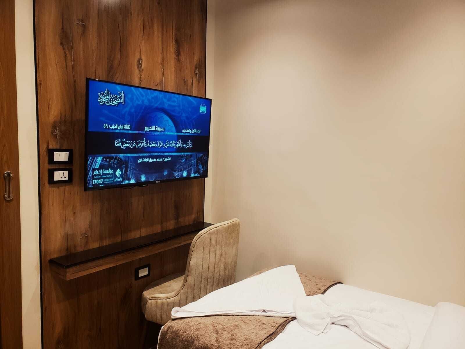 Una habitación con televisor montado en una pared de madera, una silla con cojín y una cama con manta blanca.