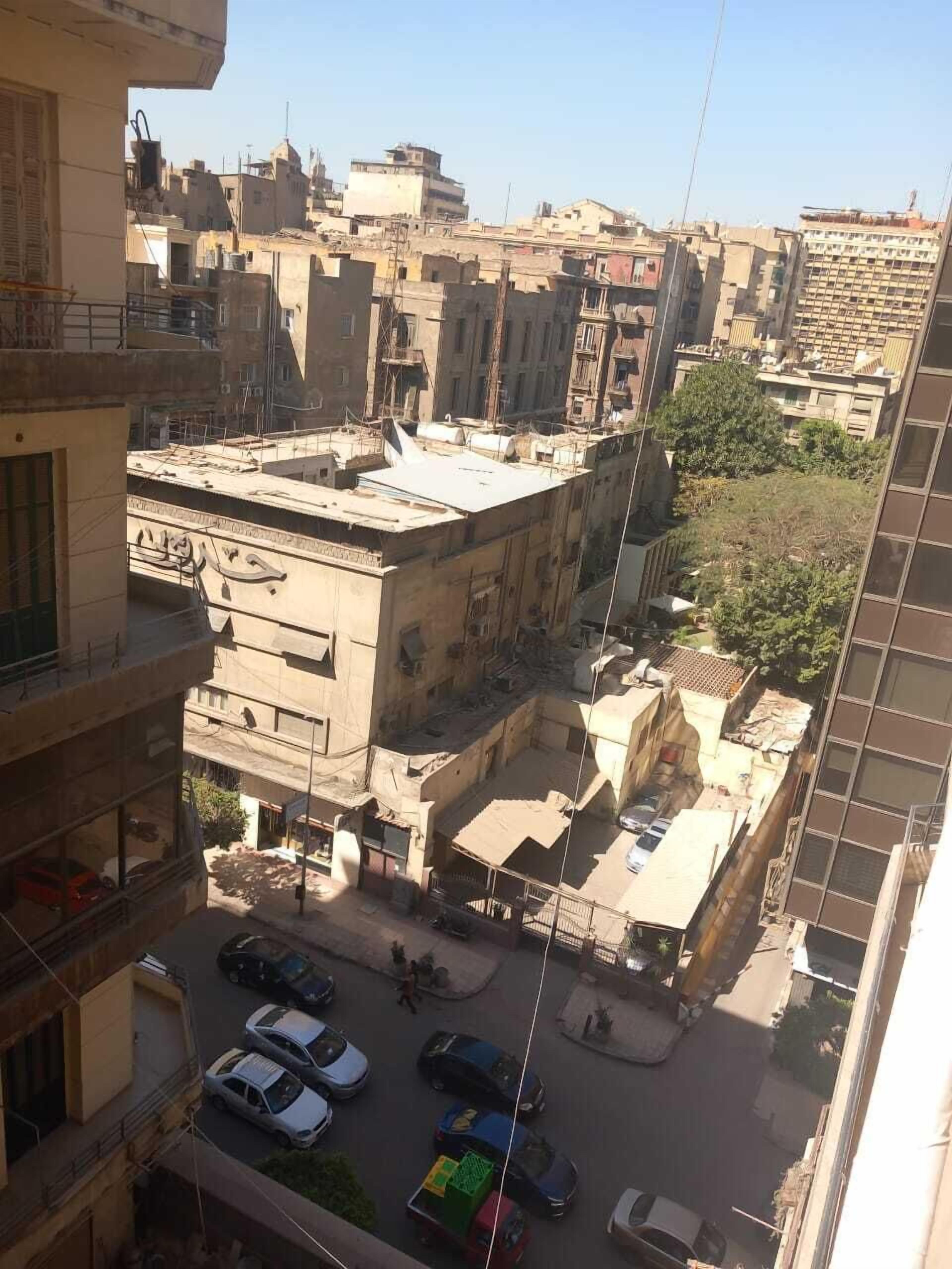 Ένα πολυώροφο κτίριο με μια αυλή, οχήματα σταθμευμένα στο δρόμο και άλλα κτίρια στο βάθος.