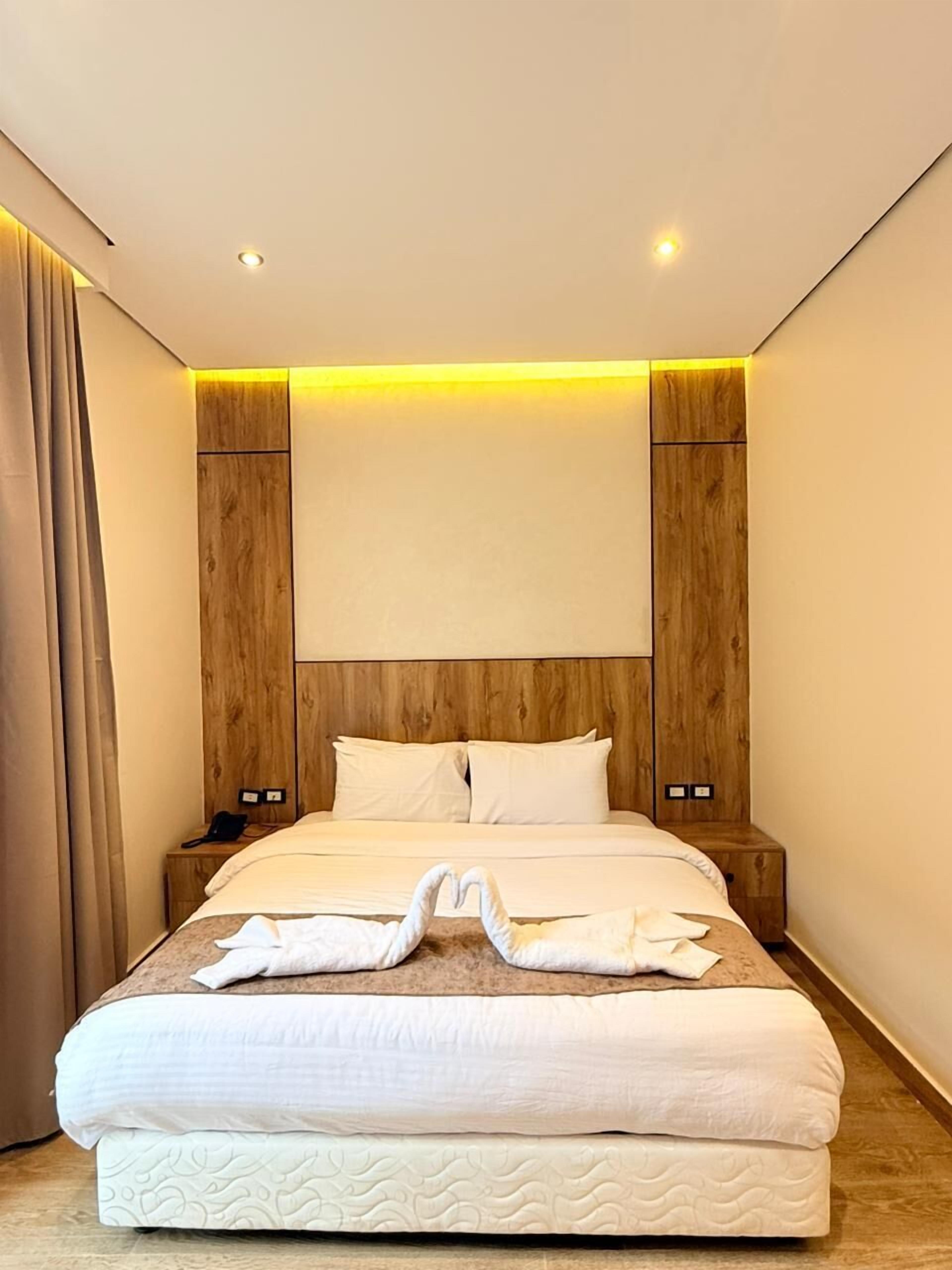 A modern hotel room with a wooden headboard, a bed with white linens, and a pair of towels arranged in a heart shape on the bed.