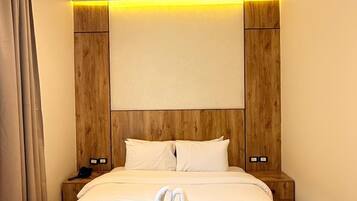 A modern hotel room with a wooden headboard, a bed with white linens, and a pair of towels arranged in a heart shape on the bed.