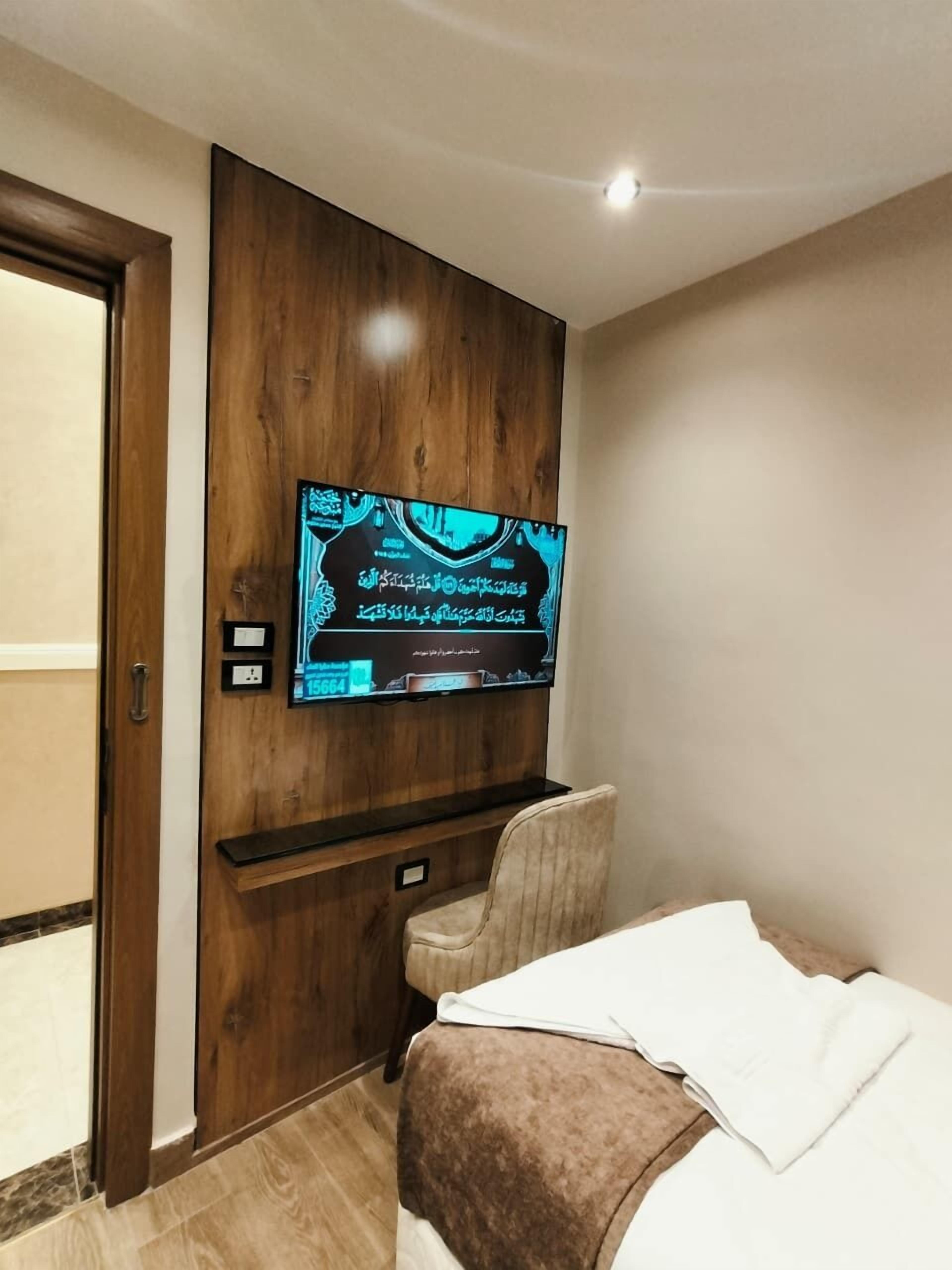 Une chambre d’hôtel avec un téléviseur à écran plat fixé sur un panneau en bois, un lit recouvert d’une couverture blanche et marron, et une chaise.