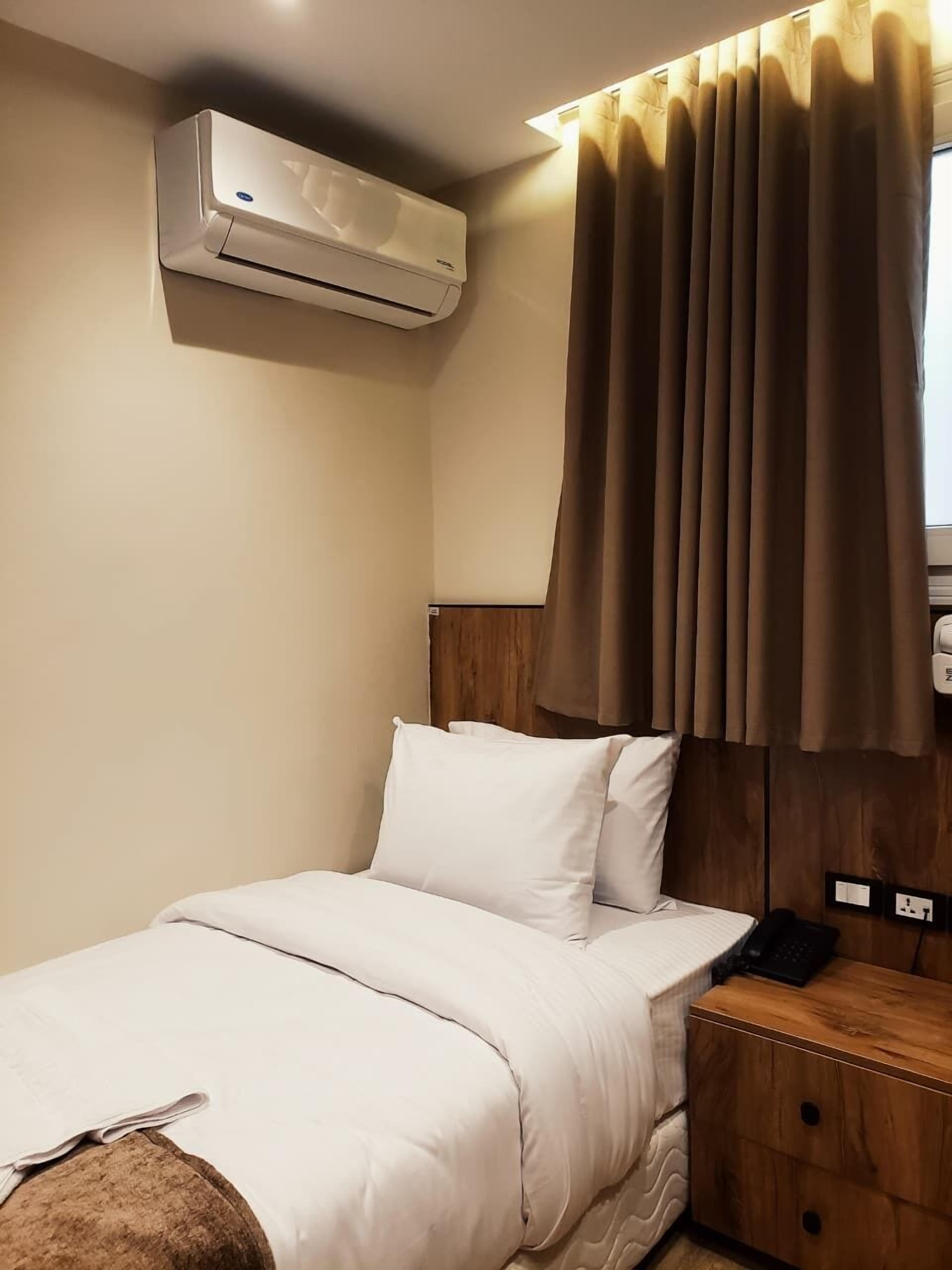 A hotel room with a bed, a bedside table with a phone, an air conditioner, and a window with curtains.
