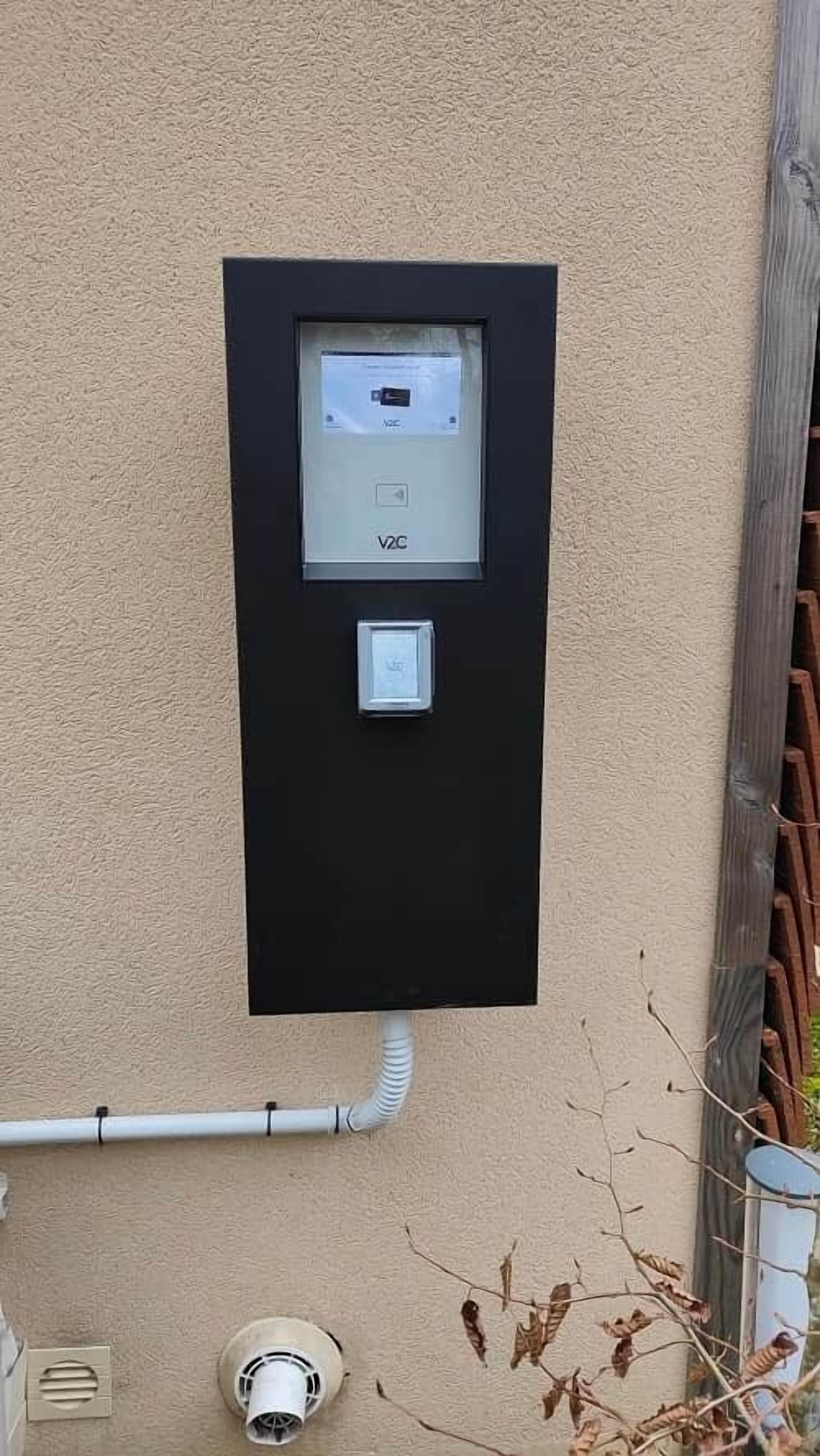 A V2C smart meter mounted on a beige wall, connected to a white pipe.