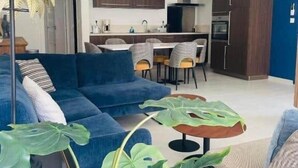 A modern living room with a blue sofa, a dining area with a kitchen in the background, and large green plants.