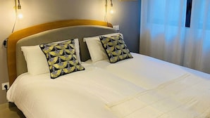 A neatly made bed with white bedding and two decorative pillows, a wall-mounted light fixture, and a window with sheer curtains.