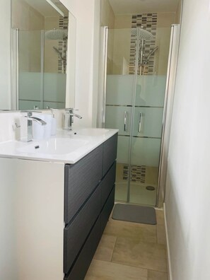 Ein modernes Badezimmer mit Duschkabine aus Glas, einem weißen Waschbecken inklusive Spiegel und einem dunklen Schrank.