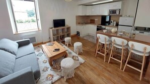 A modern living room with a sofa, coffee table, and ottomans, a dining area with chairs, and a kitchen with white cabinets and appliances. - Le logis de la lanterne (Sarlat-la-Canéda)