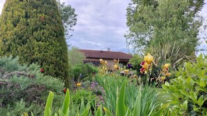 Un jardín con diversas plantas y flores, un árbol grande y un edificio con techo rojo al fondo.