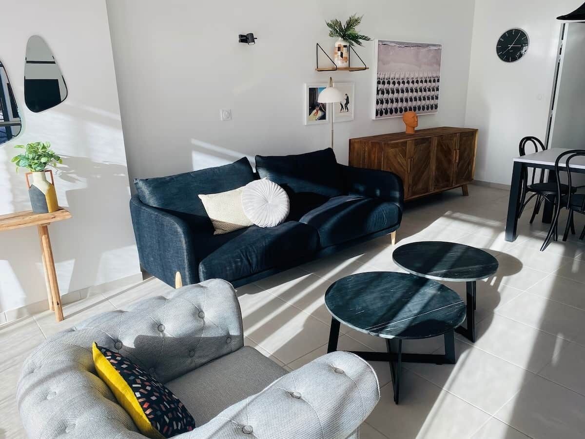 Un salon moderne avec un canapé bleu foncé, un fauteuil gris et deux petites tables rondes. On y trouve une table d’appoint en bois sur laquelle est posée une plante, une horloge murale et un tableau encadré.