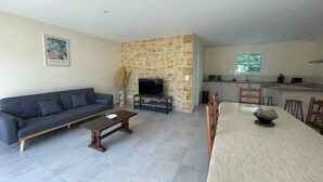 A living room with a sofa, coffee table, TV, stone wall, dining table, chairs, and a kitchen area. - House sleeps 6 pool (Sarlat-la-Canéda)