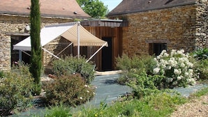 A stone building with a covered outdoor seating area, surrounded by greenery and a small garden. - Atypical pool house in a bucolic setting. (Campagnac-lès-Quercy)