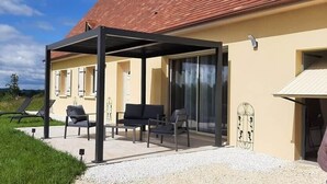 Een overdekt terras met stoelen en een tafel, grenzend aan een gebouw met een rood dak en beige muren.