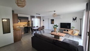 Een moderne woonkamer met een donkere bank, een houten salontafel en een televisie. Er is een zithoek met een tafel en stoelen, en een keuken in de achtergrond.