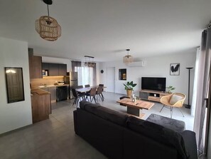 A modern living room with a dark sofa, a wooden coffee table, and a TV. There is a dining area with a table and chairs, and a kitchen in the background. - Quiet house* Valley view* Near Sarlat (Saint-André-d'Allas)