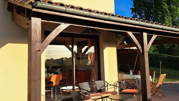 A covered outdoor seating area with chairs and tables, a pergola structure, and a view of the interior through glass doors. - Villa*Piscine*Proche Sarlat (Proissans)