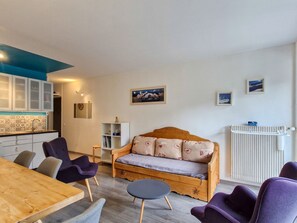 A cozy living room with a wooden sofa, a dining table, and purple chairs. There is a kitchen area with cabinets and a patterned backsplash. - Apartment in resort center, slope view - 8 guests (Flaine)