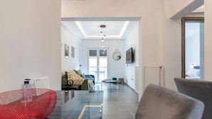 A modern living room with a glass coffee table, a sofa, and a red chair. There is a television mounted on the wall and a window with a view of the city. - Daedalus Minimal 1bedroom Bliss (Athina)