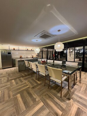 A modern kitchen with a dining area, featuring a herringbone wood floor, a glass dining table, wicker chairs, and pendant lighting.
