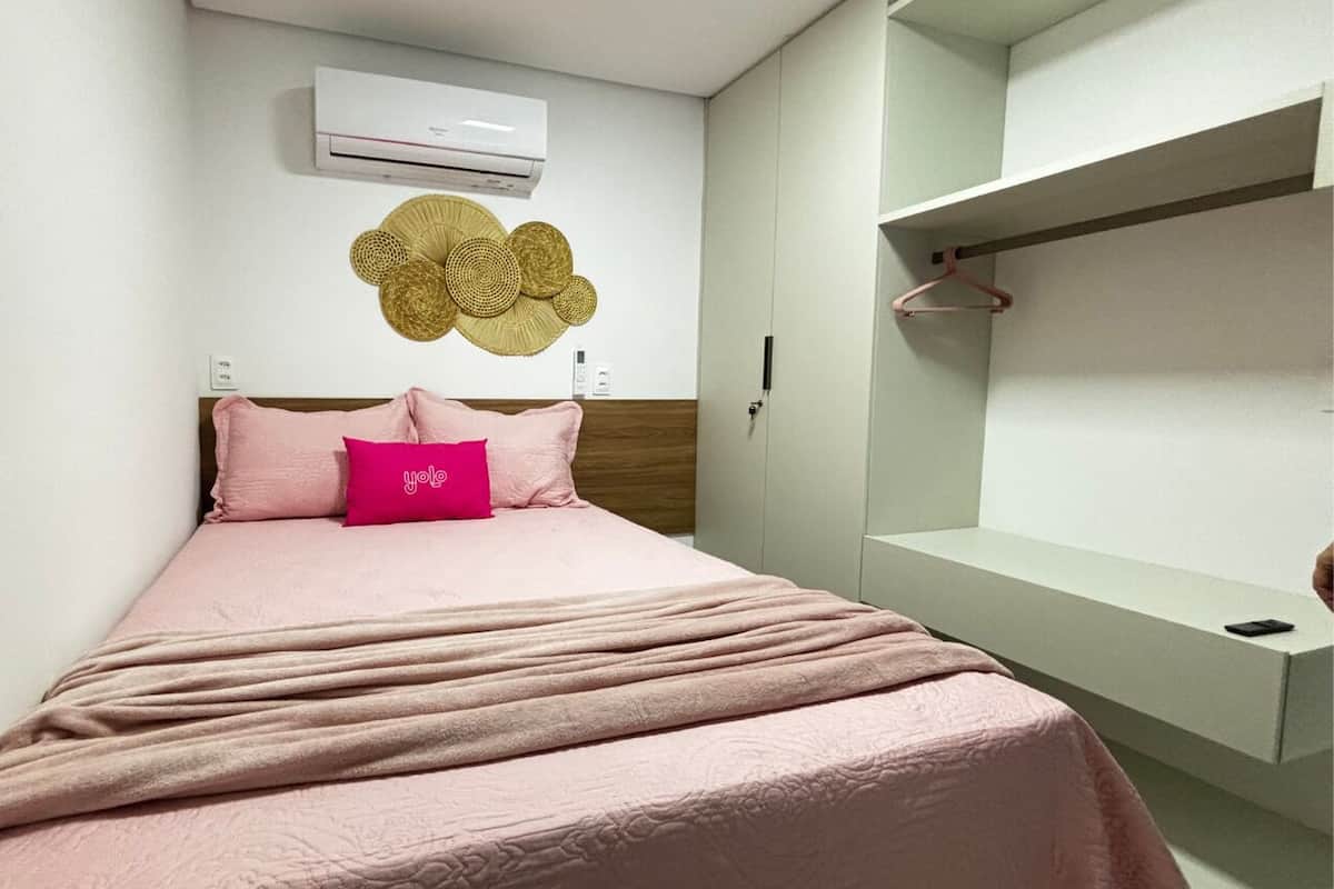 A bedroom with a bed, a wall-mounted air conditioner, a decorative wall piece, and a wardrobe with a hanger.