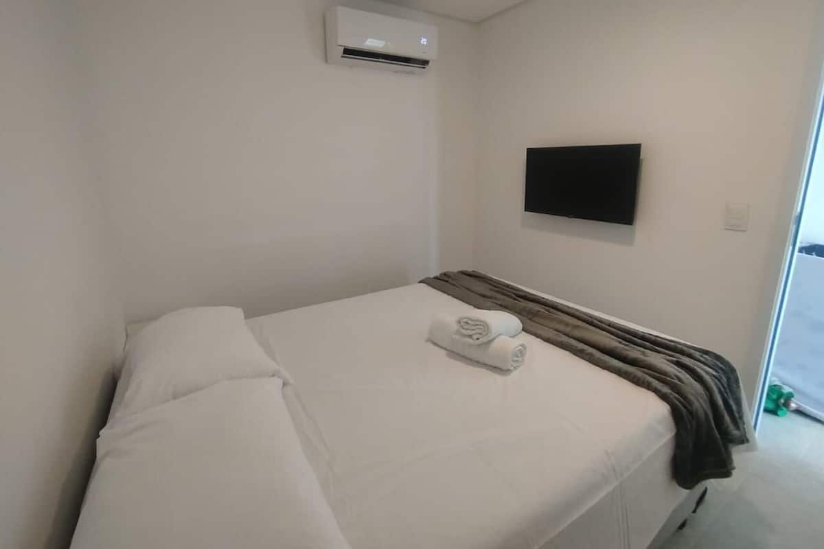 A hotel room with a bed, a flat-screen TV mounted on the wall, and an air conditioning unit above the bed.