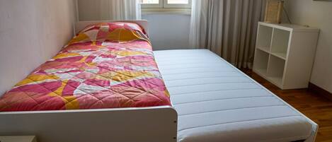 A single bed with a quilt, a bedside table, a lamp, and a window with curtains.