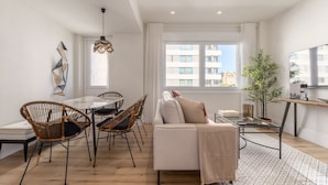 Een moderne woonkamer met een eetgedeelte, met een bank, stoelen, een eettafel en een groot raam met uitzicht op gebouwen.