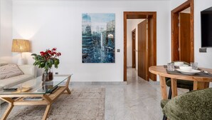 Un soggiorno moderno con un tavolino in vetro, un divano, un'area pranzo con tavolo e sedie in legno e un dipinto con vista sulla città.
