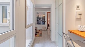 Un corridoio con una porta in vetro, un quadro che raffigura una veduta cittadina, un pavimento in marmo e una mensola in legno con una borsa bianca.