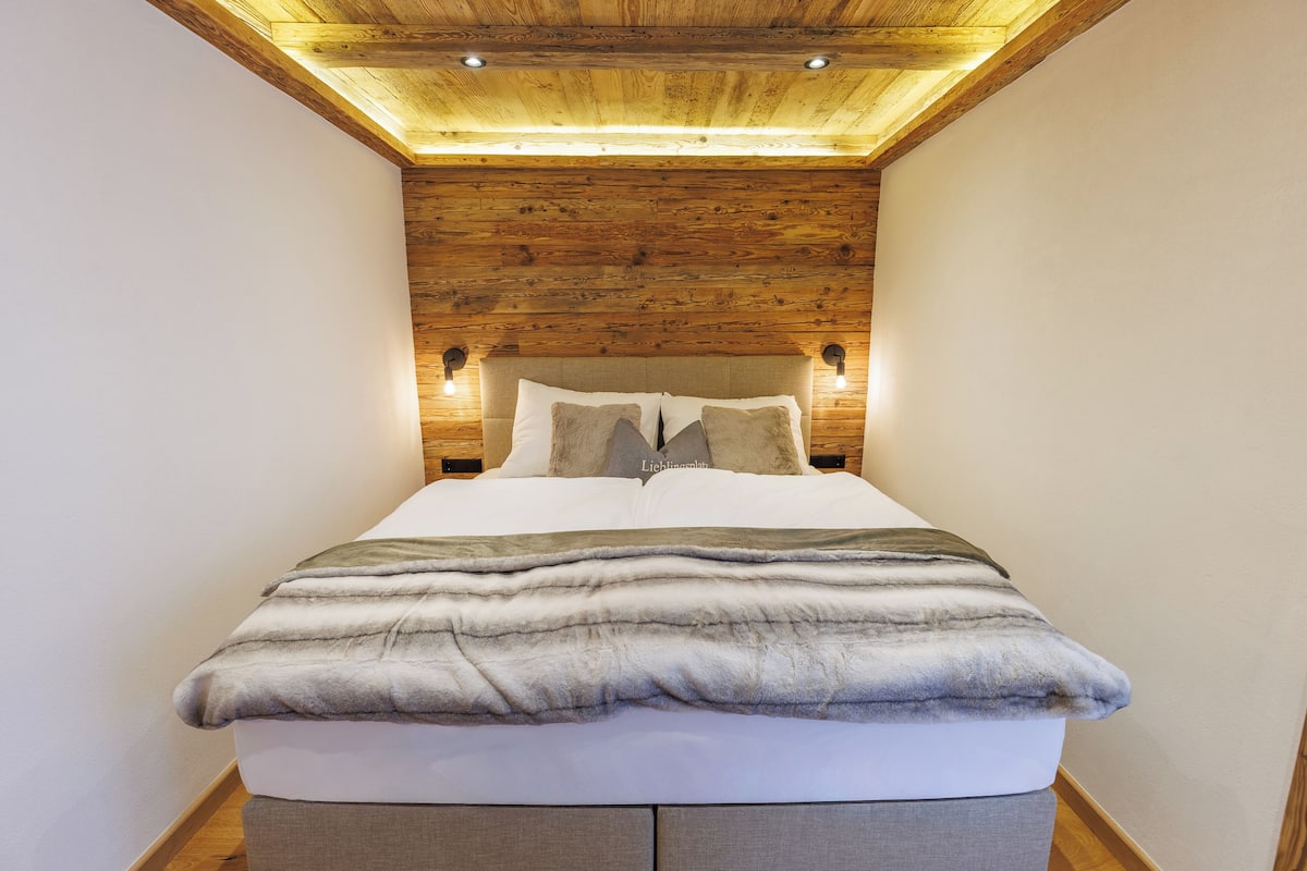 A bedroom with a wooden ceiling, a bed with a grey blanket, and two wall-mounted lamps.