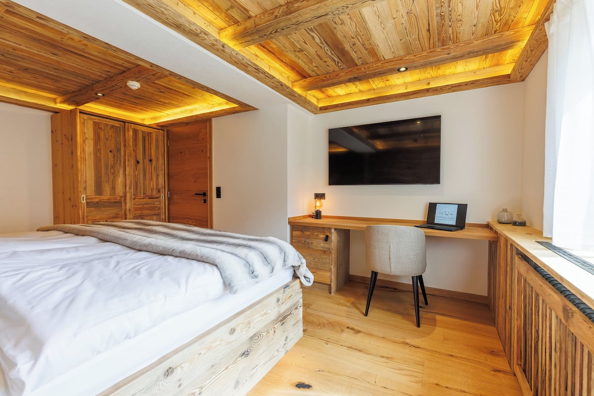 Um quarto com cama de madeira, escrivaninha com cadeira, televisão e teto de madeira.